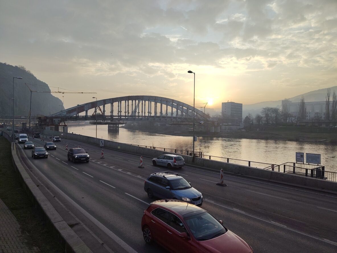 Auta projíždějící na nábřeží, řeka Labe a slunce vycházející za rekonstruovaným Benešovým mostem v Ústí nad Labem